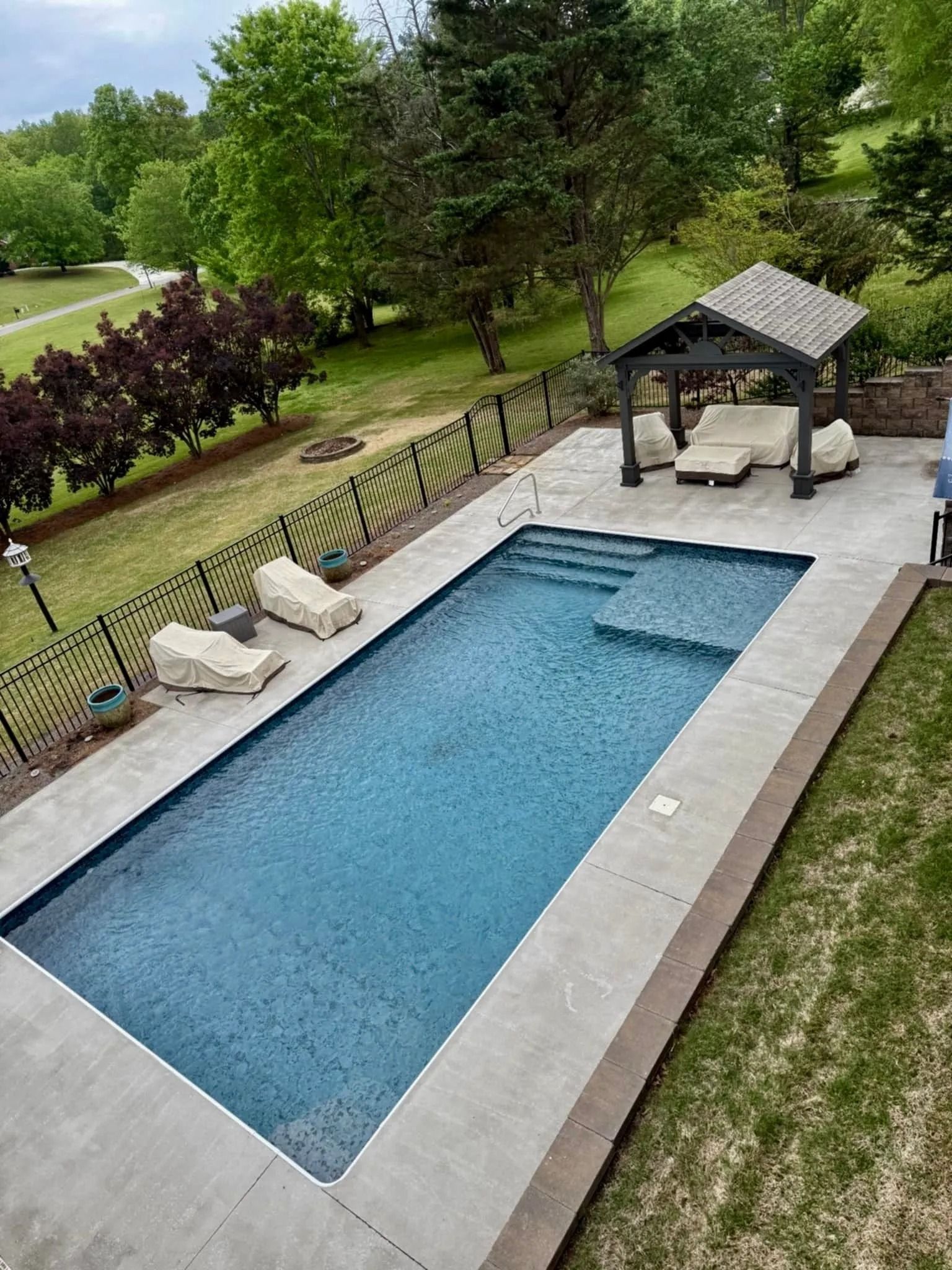 Backyard with a rectangular blue swimming pool, lounge chairs, and a shaded cabana on a grassy lawn