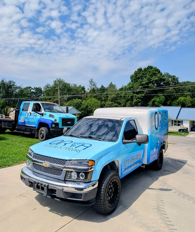 Blue tow truck parked on gravel under trees, with a trailer behind it.