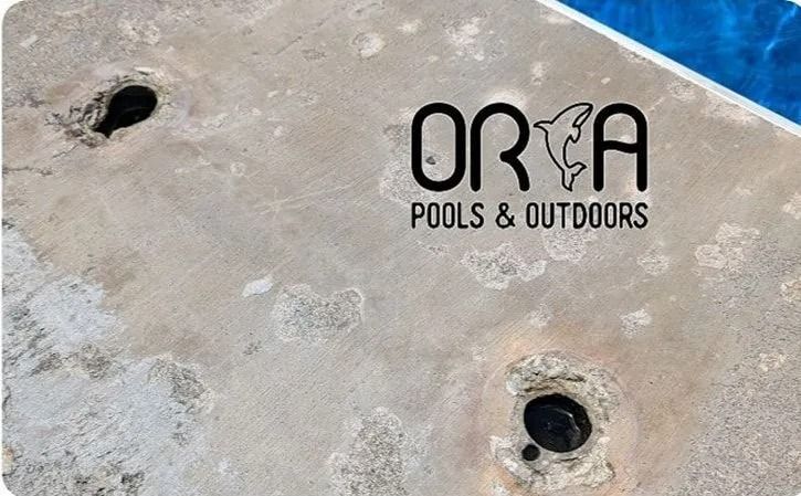 Concrete pool surface with two dark drain openings and ORA Pools & Outdoors logo.