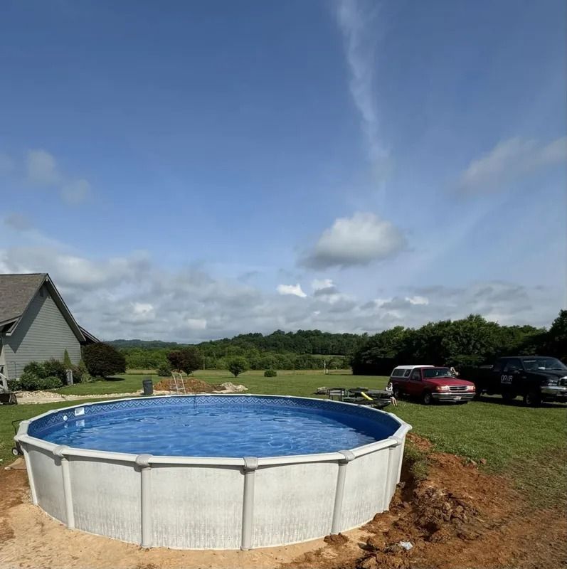 Above-ground pool in a grassy yard beside a house, with cars and trees under a blue sky.