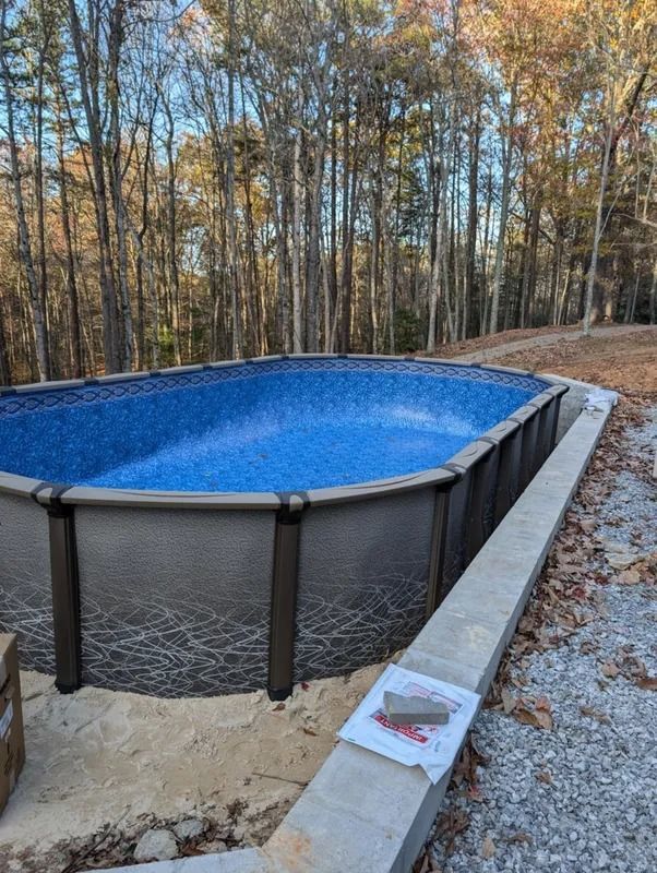 Above-ground pool with blue water in a wooded backyard, surrounded by a black safety fence