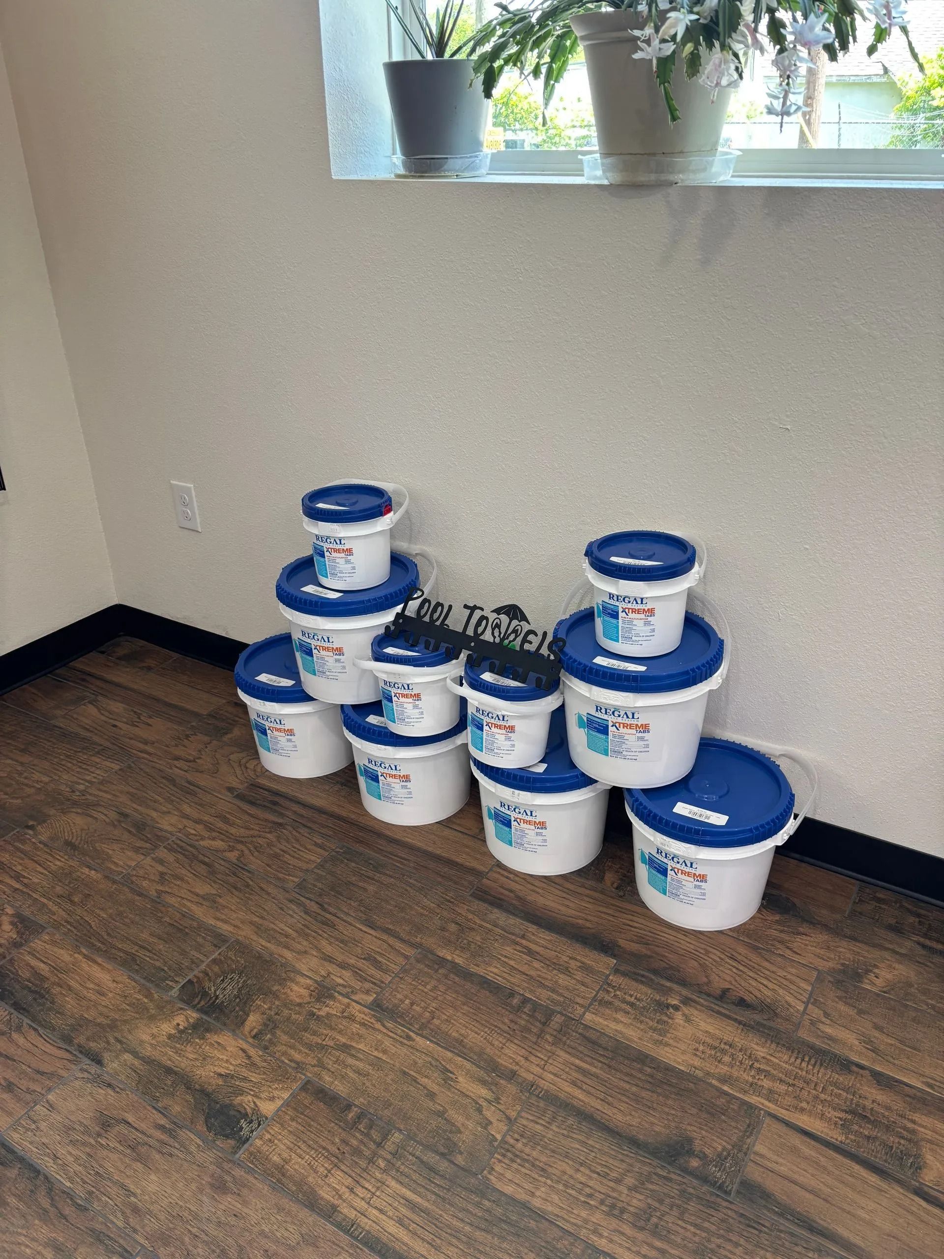 Stacked white-and-blue paint buckets in a room corner beneath a window with potted plants.