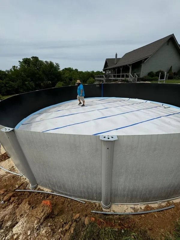 Person standing inside a large circular above-ground pool with a white liner and blue support straps, outdoors.