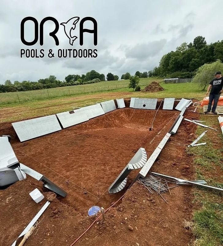 Partially built backyard pool shell with construction materials on red dirt, under cloudy sky