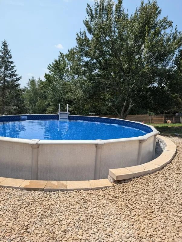 Round above-ground pool with blue water in a gravel backyard, surrounded by trees and a tan deck.