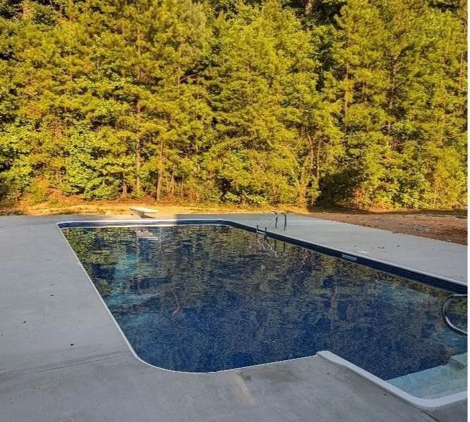 Backyard swimming pool with blue water beside a concrete deck and tall green trees