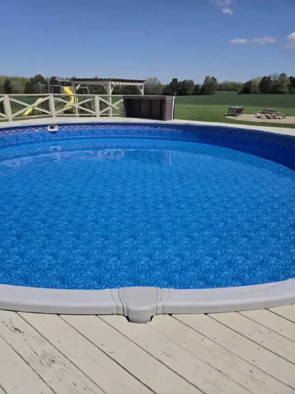 Round blue swimming pool on a wooden deck with a grassy field and white fence in the background