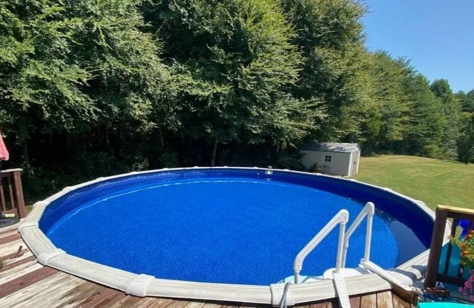 Round blue backyard swimming pool surrounded by wooden deck and trees, with ladder in the foreground