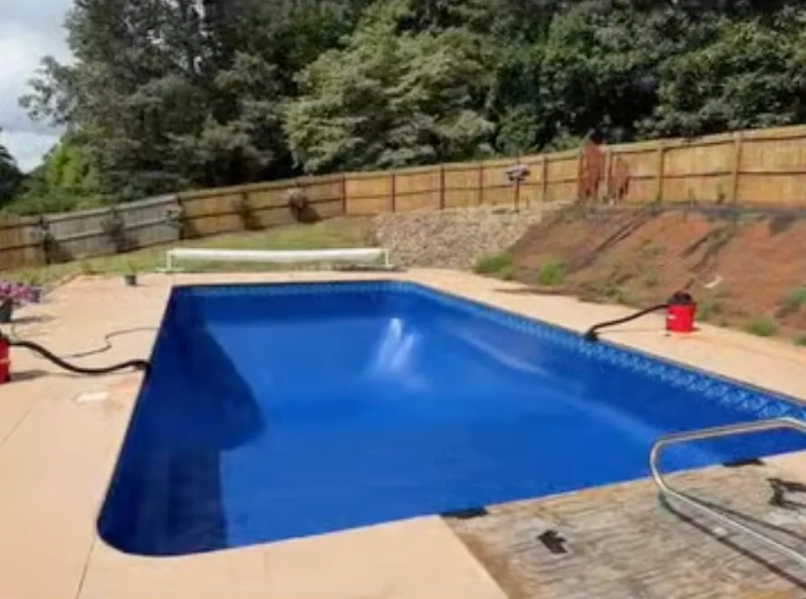 Backyard swimming pool with blue water, tan deck, and a wooden fence along a hillside.