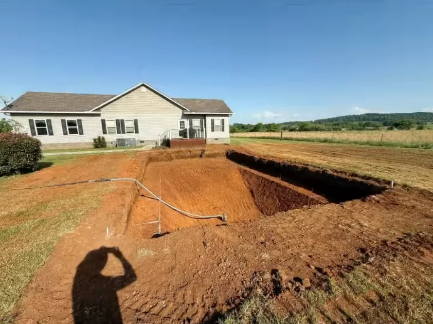 Large backyard excavation beside a beige house, with a trench and open field under a clear blue sky.