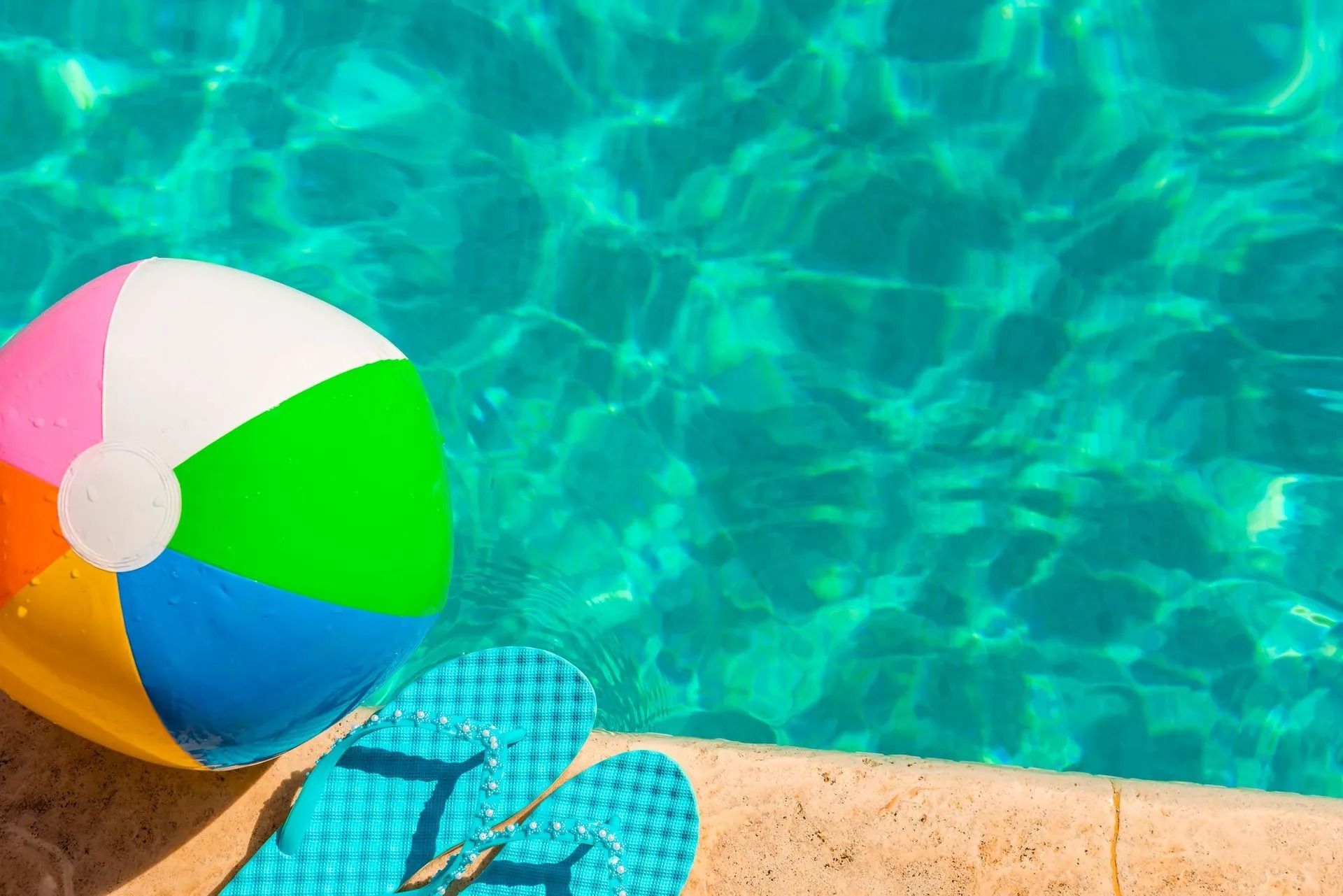 Colorful beach ball and blue flip-flops beside a turquoise swimming pool