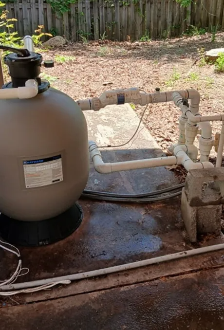 Pool filtration equipment with pipes and a beige tank on a concrete pad in a backyard