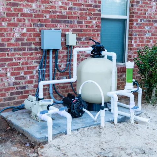 Outdoor pool filtration equipment with white pipes, pumps, and tanks against a brick wall