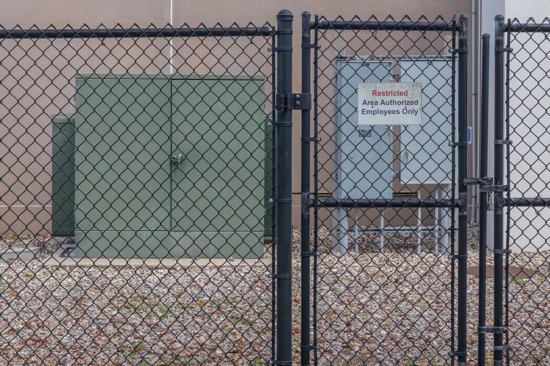 Chain-link fence with warning sign, green utility box, and table in a gravel lot.