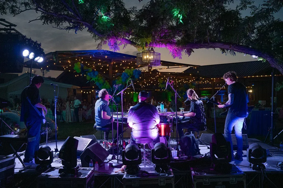 Live band performing outdoors at night under trees with purple stage lights