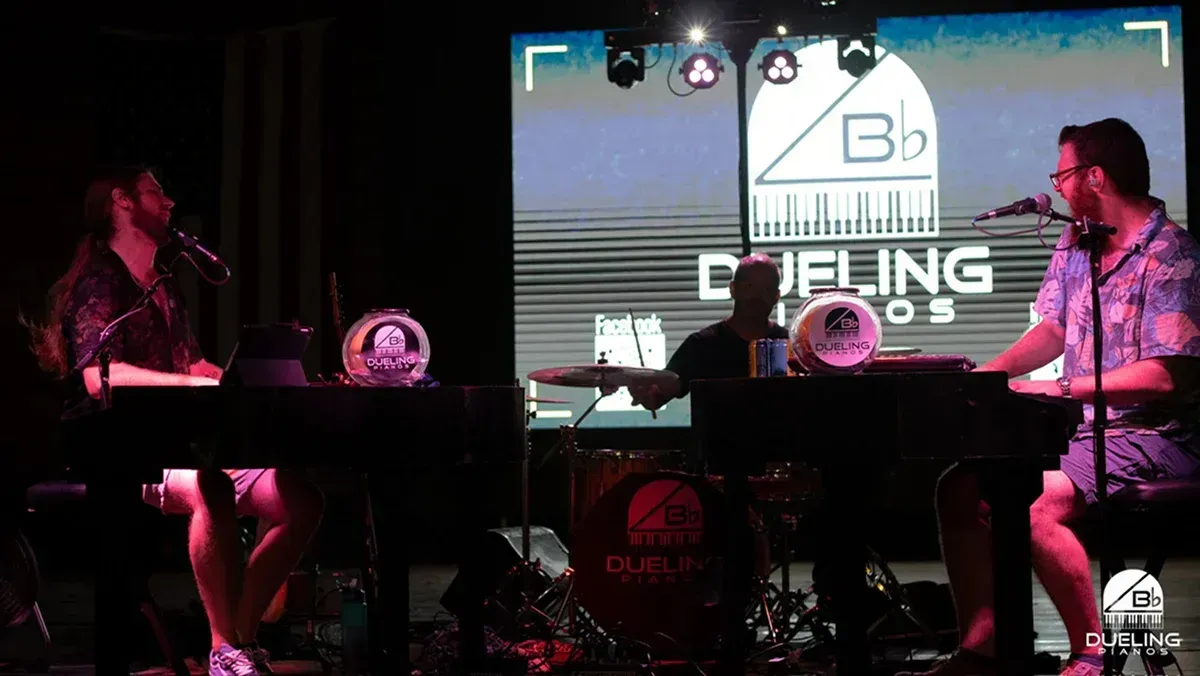 Two musicians seated at keyboards on a dim stage with pink lighting and a “Defining...” sign behind them