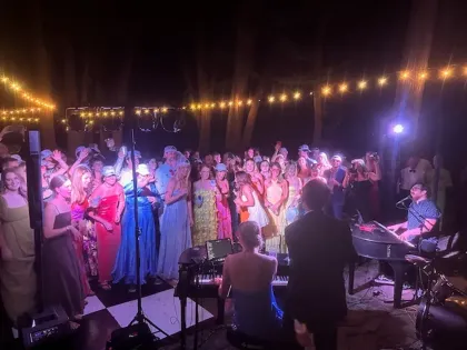 Choir performing outdoors at night under string lights, with a conductor and musicians on stage