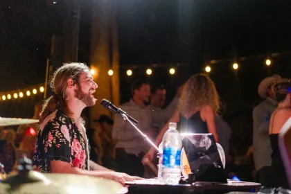 Musician singing into a microphone at a keyboard during an outdoor nighttime performance with string lights