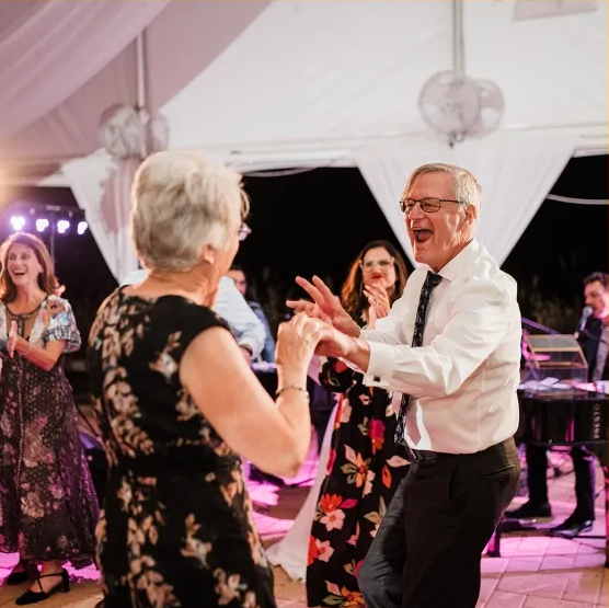 Guests dancing at a tented party, with a smiling man in a white shirt and black tie in the foreground