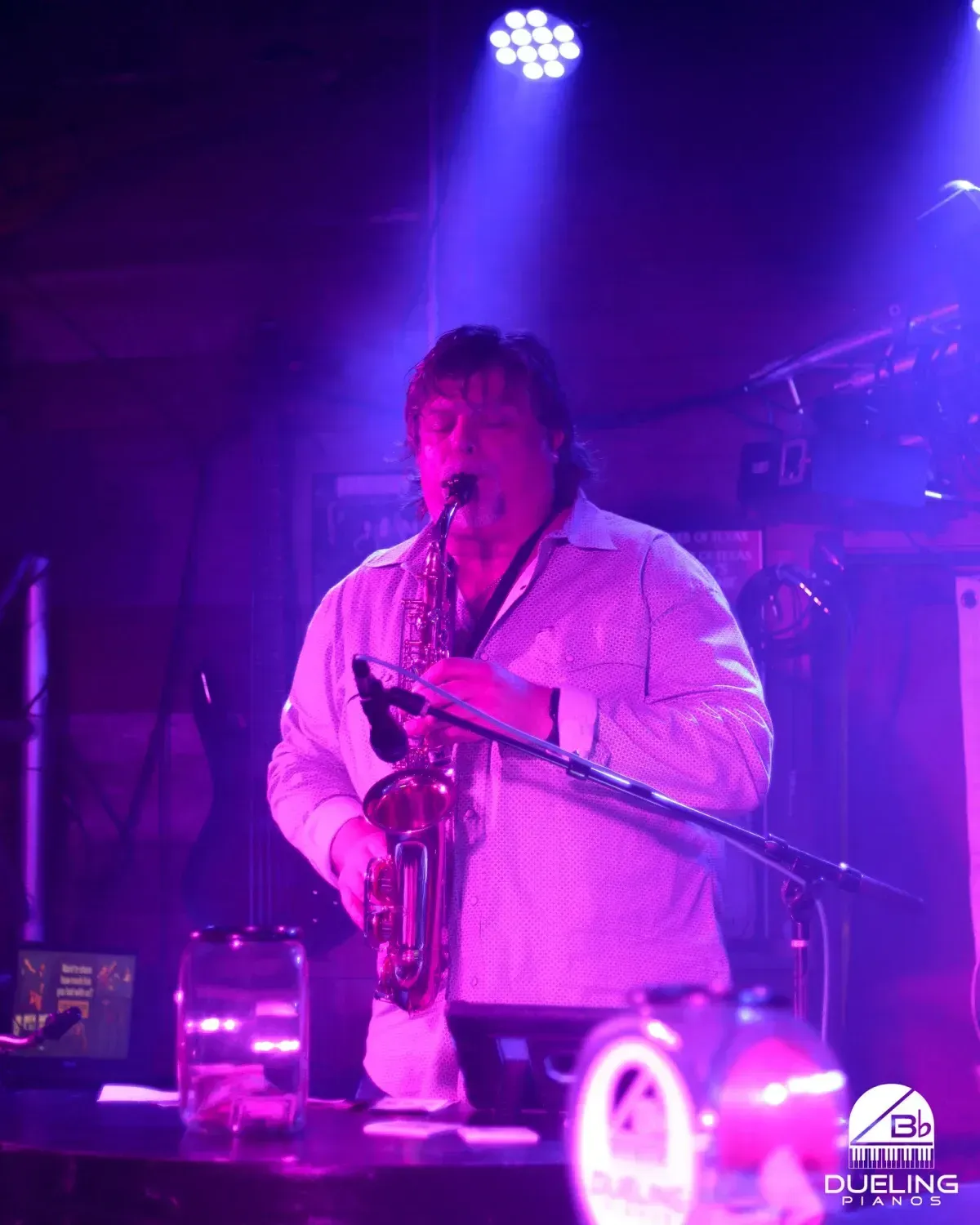 Musician playing saxophone on a purple-lit stage with microphone and drinks in the foreground