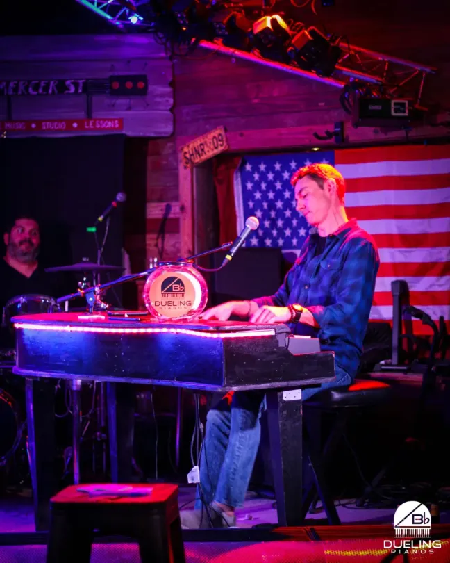 Musician at keyboard on neon-lit stage with American flag backdrop, singing into a microphone