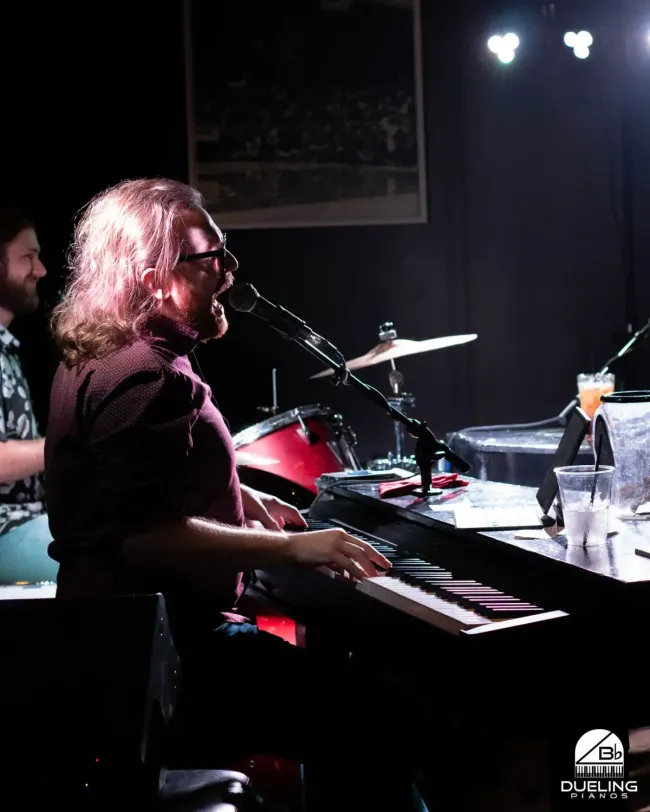 Musician singing and playing keyboard on a dimly lit stage with drum kit behind him