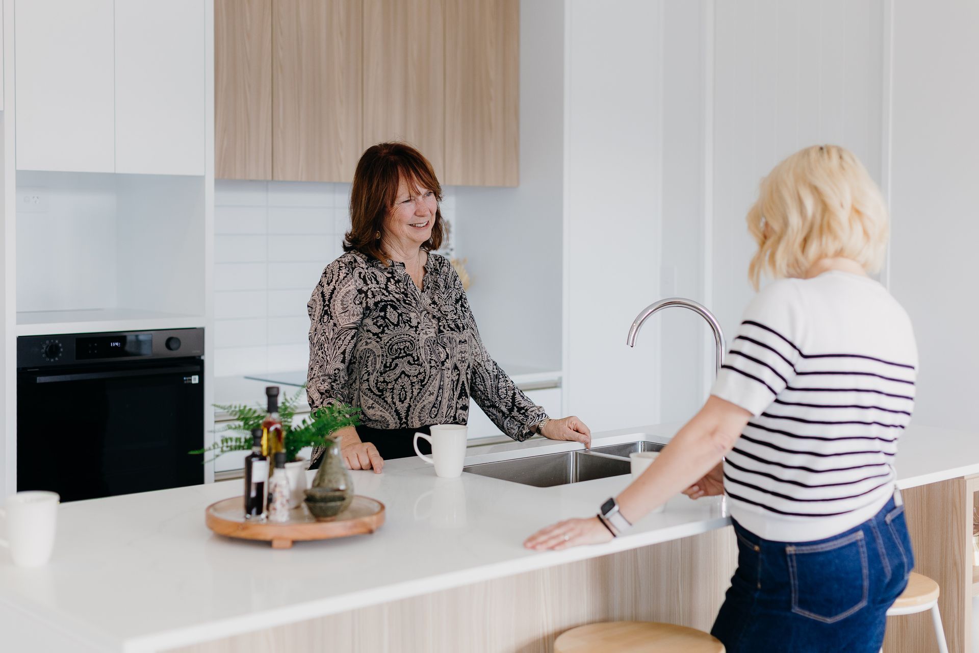 Two women at a kitchen island, one smiling. Kitchen has white counters, cabinets, and appliances.