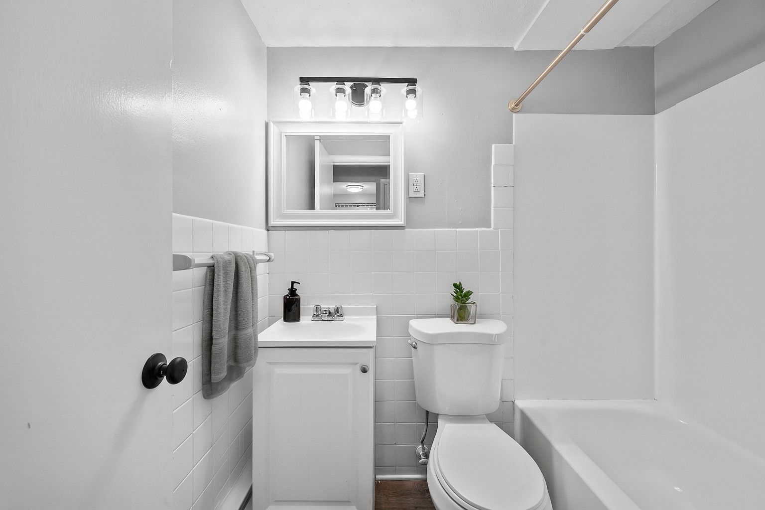 Small, white bathroom with a toilet, vanity, and tub. Gray towels hang on a wall.