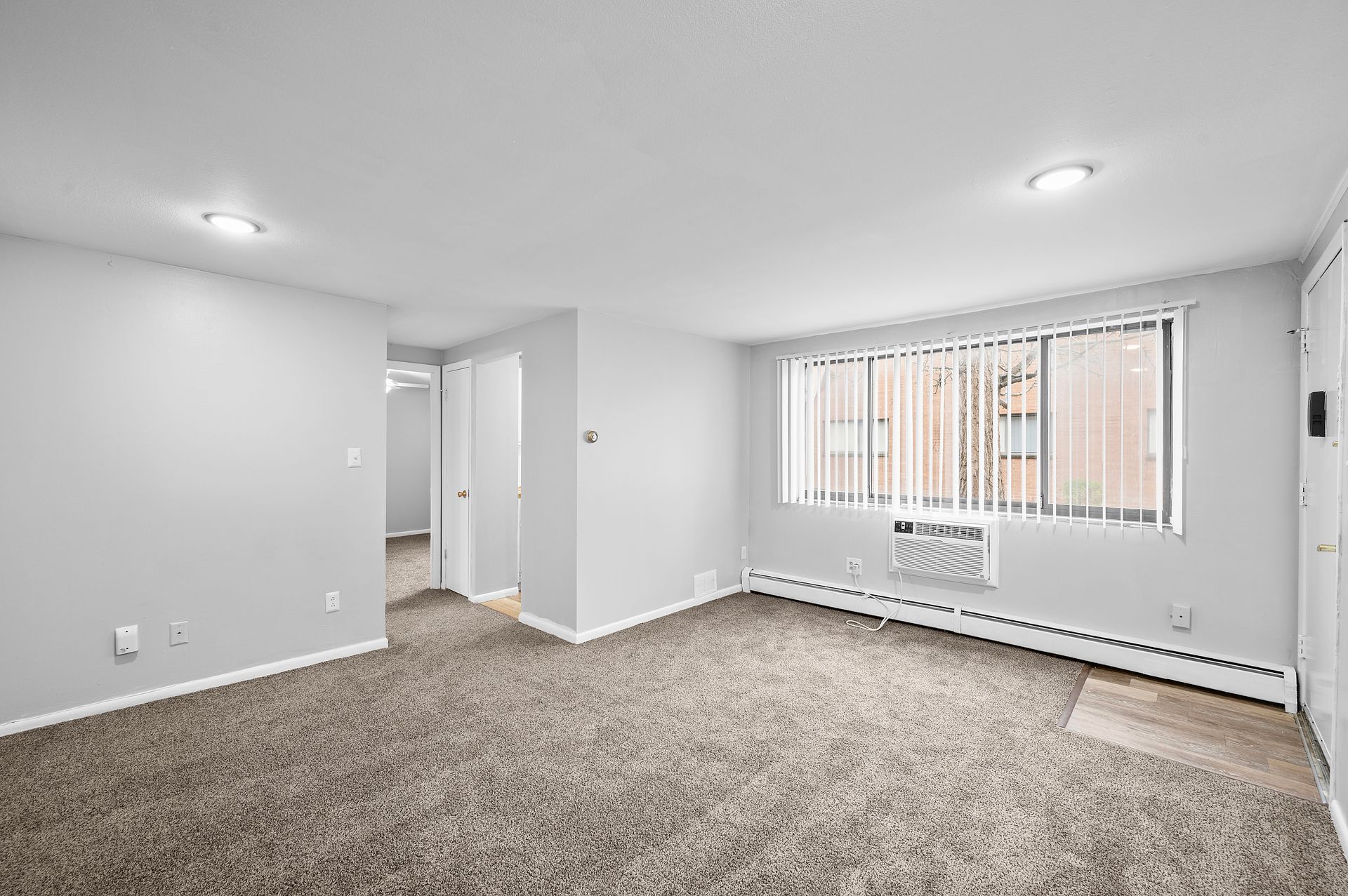 Empty apartment living room with carpet, window with blinds, and an AC unit.
