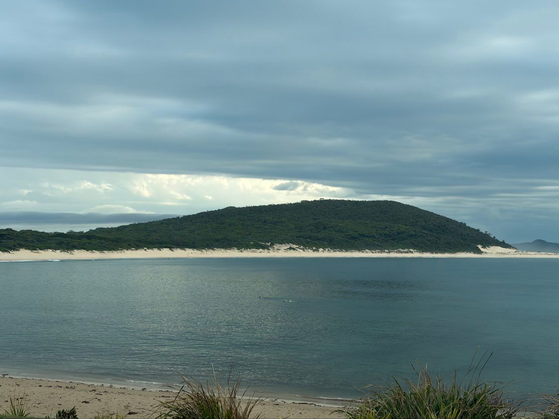 Island with dense green trees on top, sandy shore, and calm blue water under a cloudy sky.