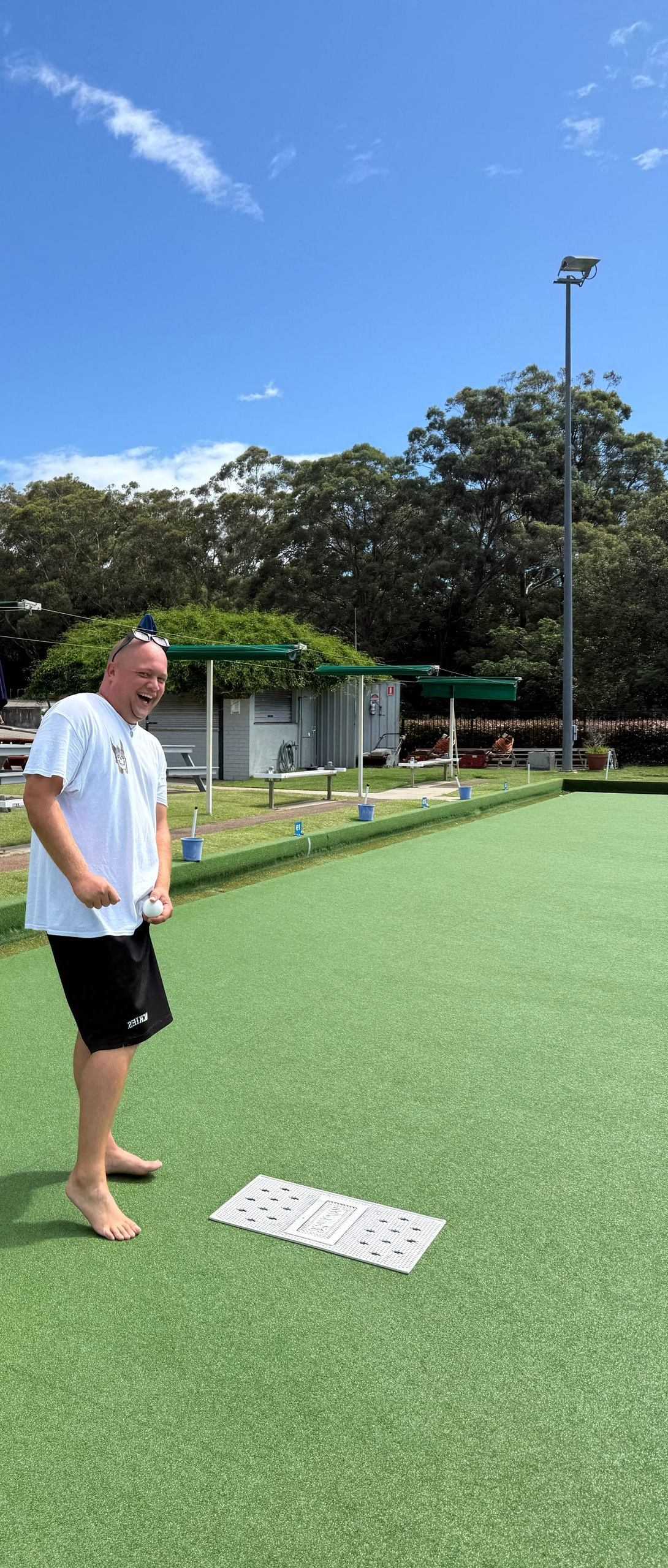 Man stands barefoot on a green lawn, smiling. He wears a white shirt and black shorts. Blue sky and trees in background.