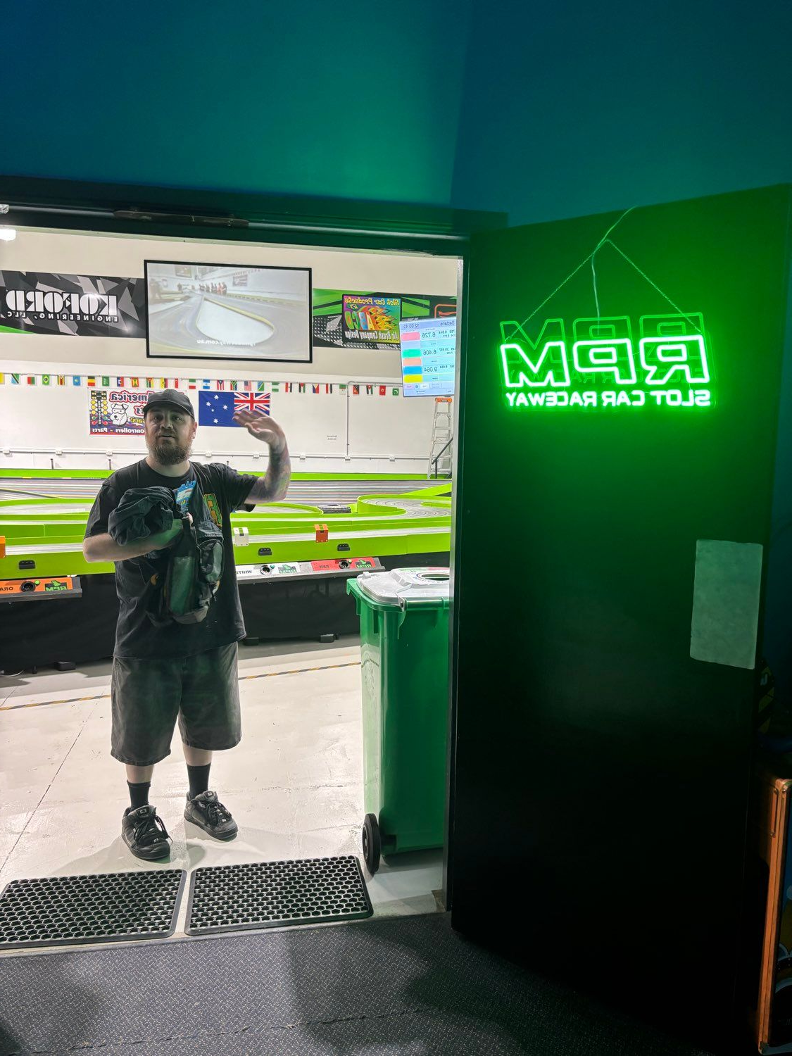 Man waves from doorway of RPM Slot Car Racing. Neon green sign and track visible.