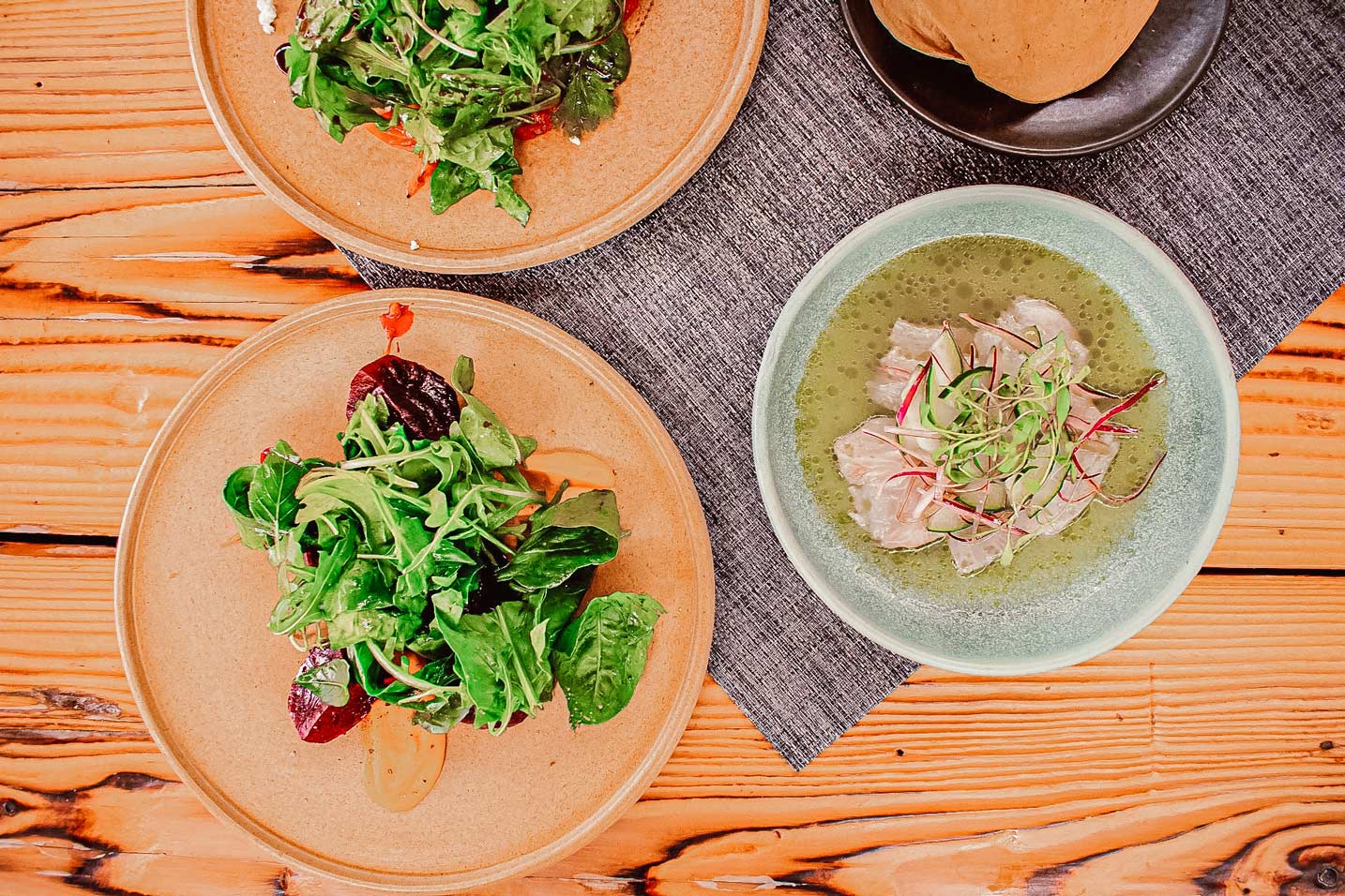 Three plates of salad and two bowls of soup are on a wooden table.