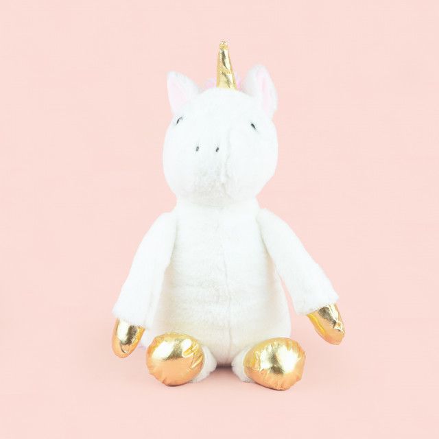 A white stuffed unicorn with gold feet and legs is sitting on a pink surface.