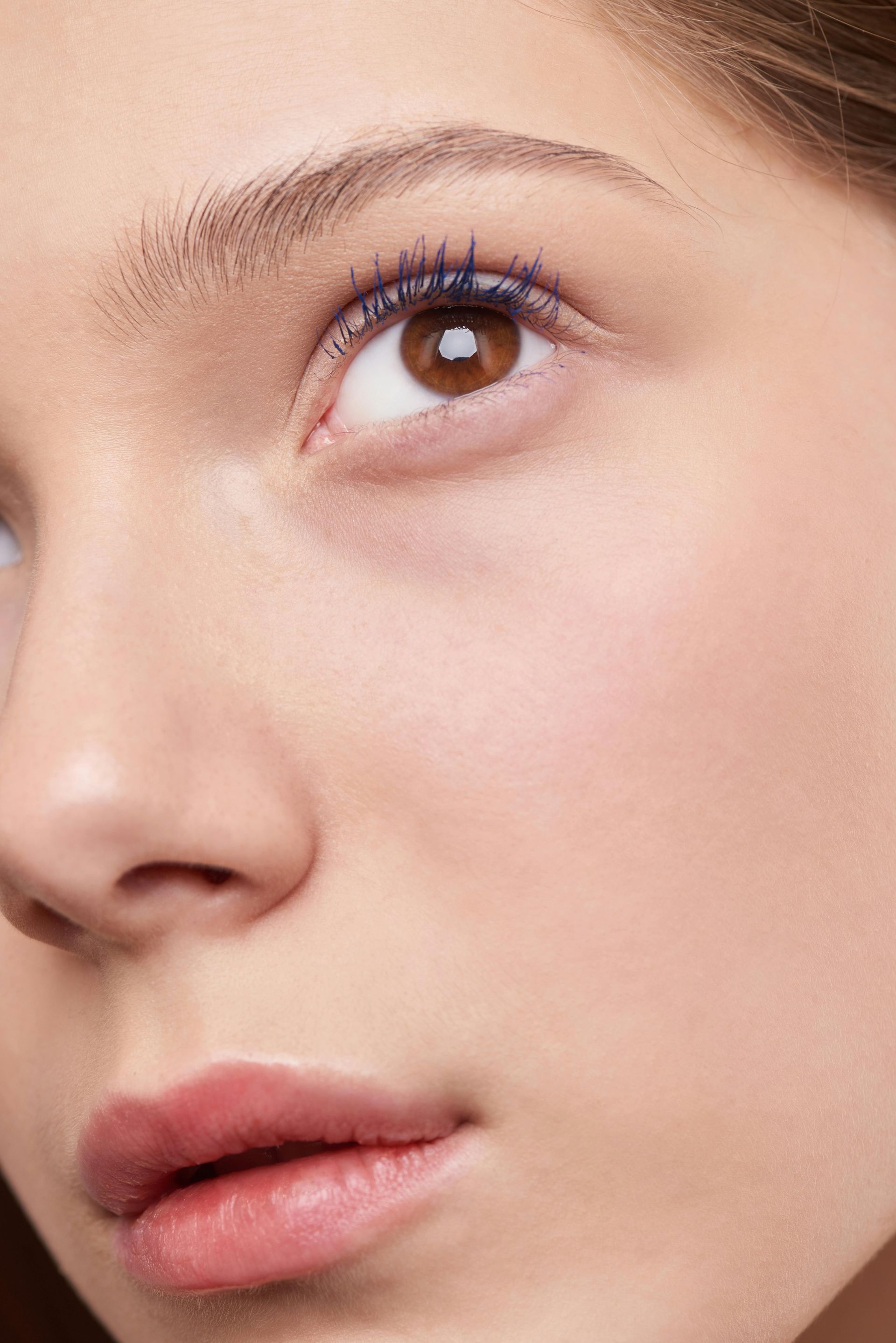 Close-up of a person with brown eyes, applied blue mascara, and mauve lipstick, looking up.
