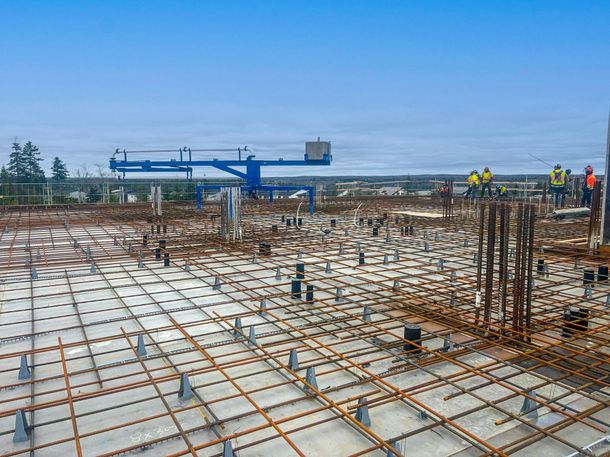 Construction site with rebar grid, blue crane, and workers on a concrete slab under a cloudy sky
