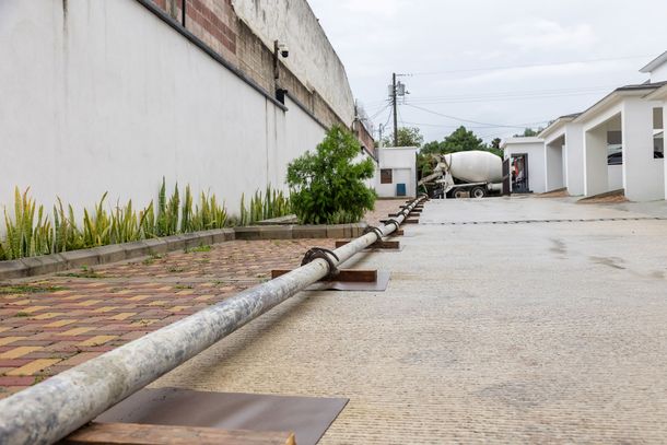 Long metal pole on a paved outdoor walkway beside a wall and white buildings under a cloudy sky