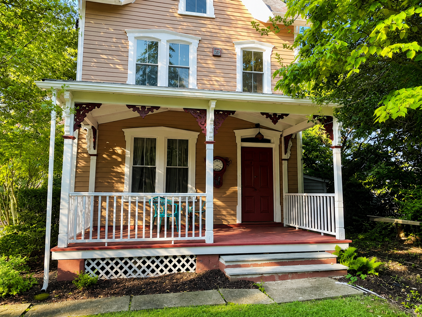 Small white house with a covered front porch, red door, and tan shingled roof surrounded by trees.