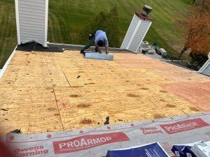 Worker installing roofing plywood on a house roof, with chimney vents and grassy yard below.