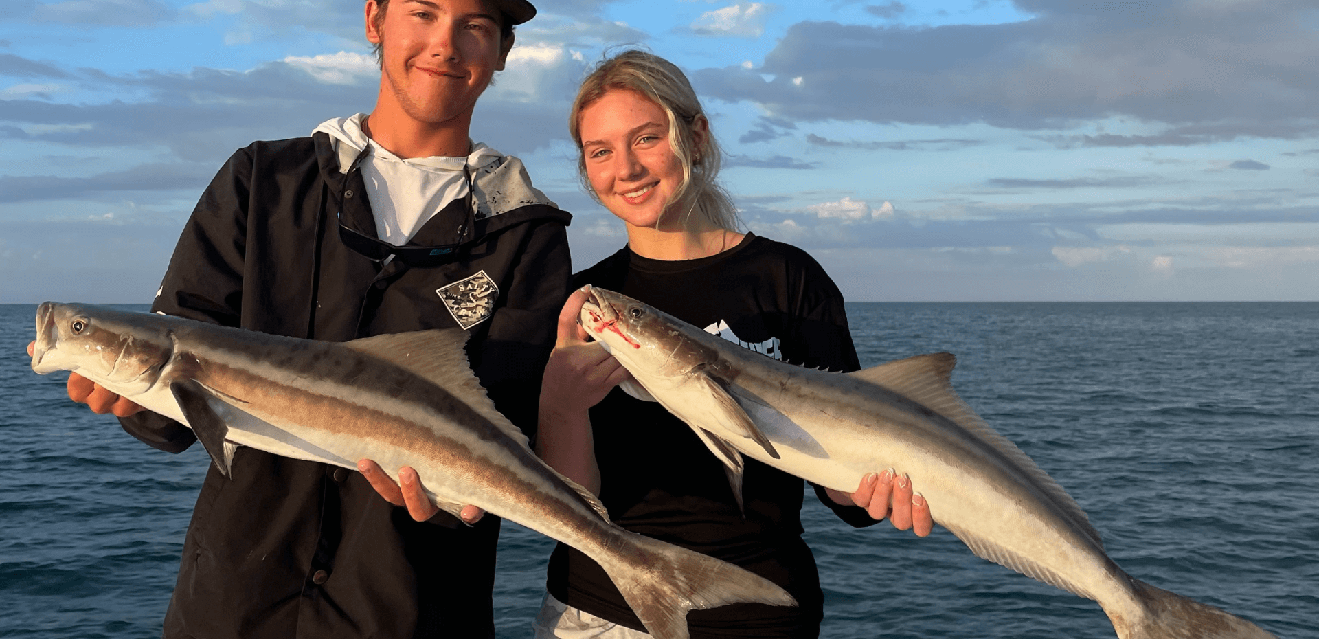 A man and a woman are holding two fish in front of the ocean.