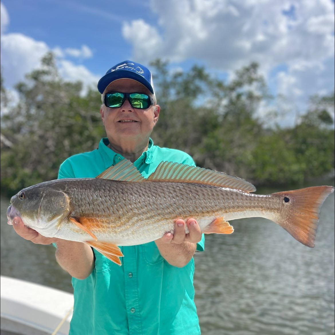 A man wearing sunglasses is holding a large red drum fish in his hands.