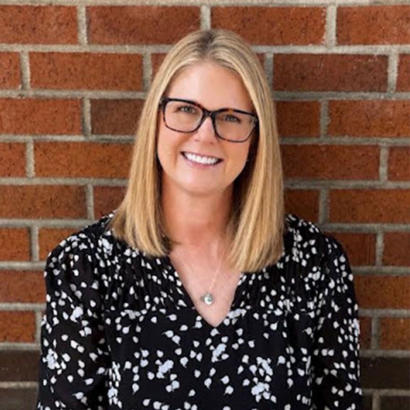 Smiling woman with glasses in a black patterned blouse standing against a brick wall