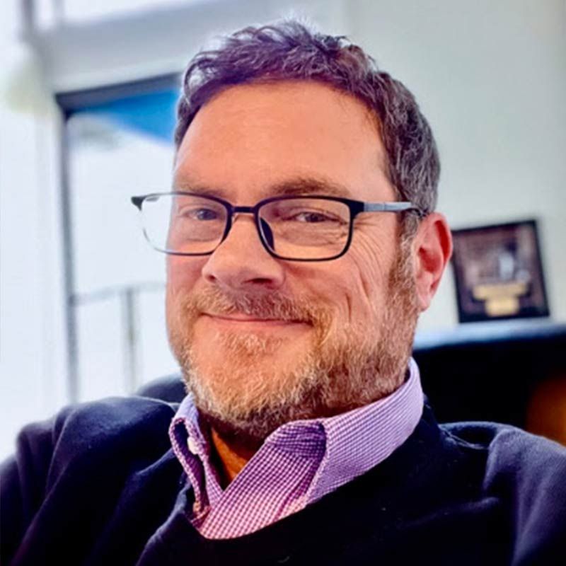 Smiling man with glasses and gray beard in a purple shirt and dark sweater, seated indoors