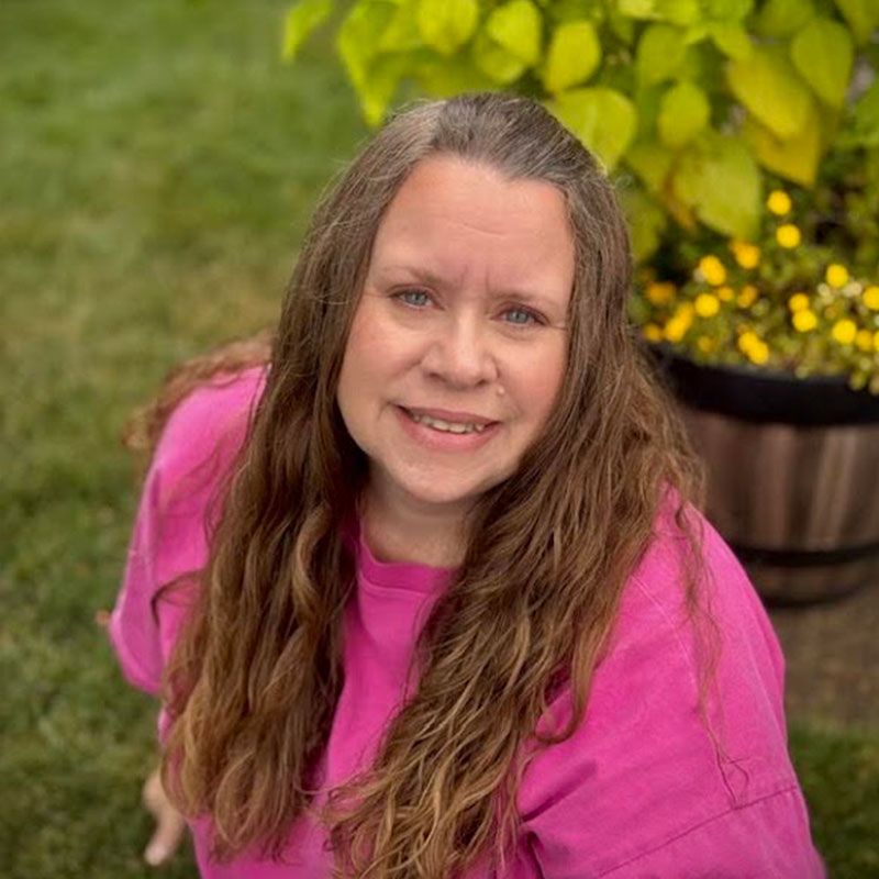 Woman in a bright pink shirt smiling outdoors beside yellow flowers and green grass