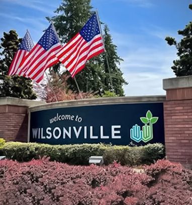 Wilsonville welcome sign with American flags, brick wall, and pink shrubs under a blue sky