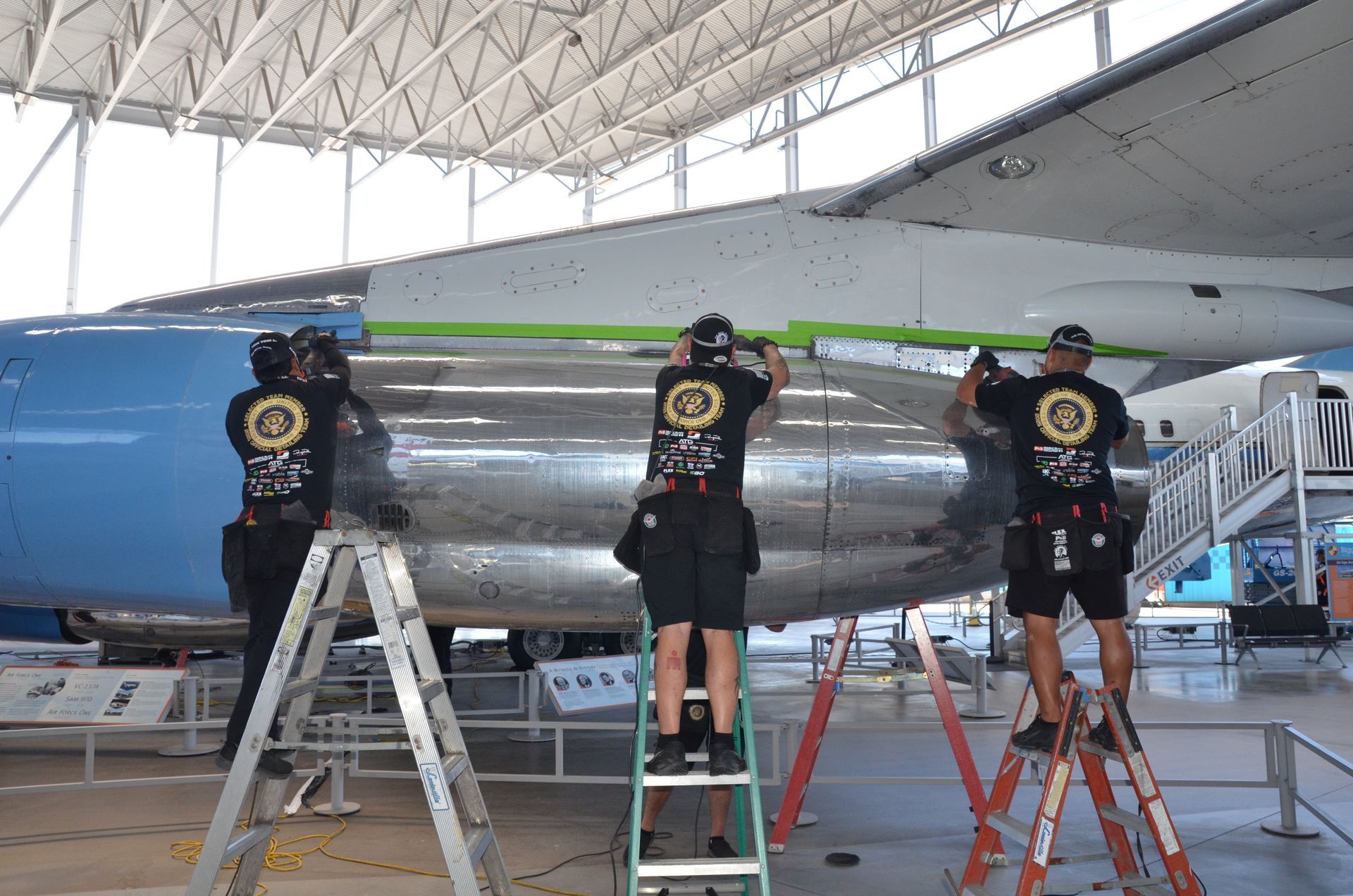 Four people work on the side of a large blue and silver airplane, using ladders, inside a hangar.