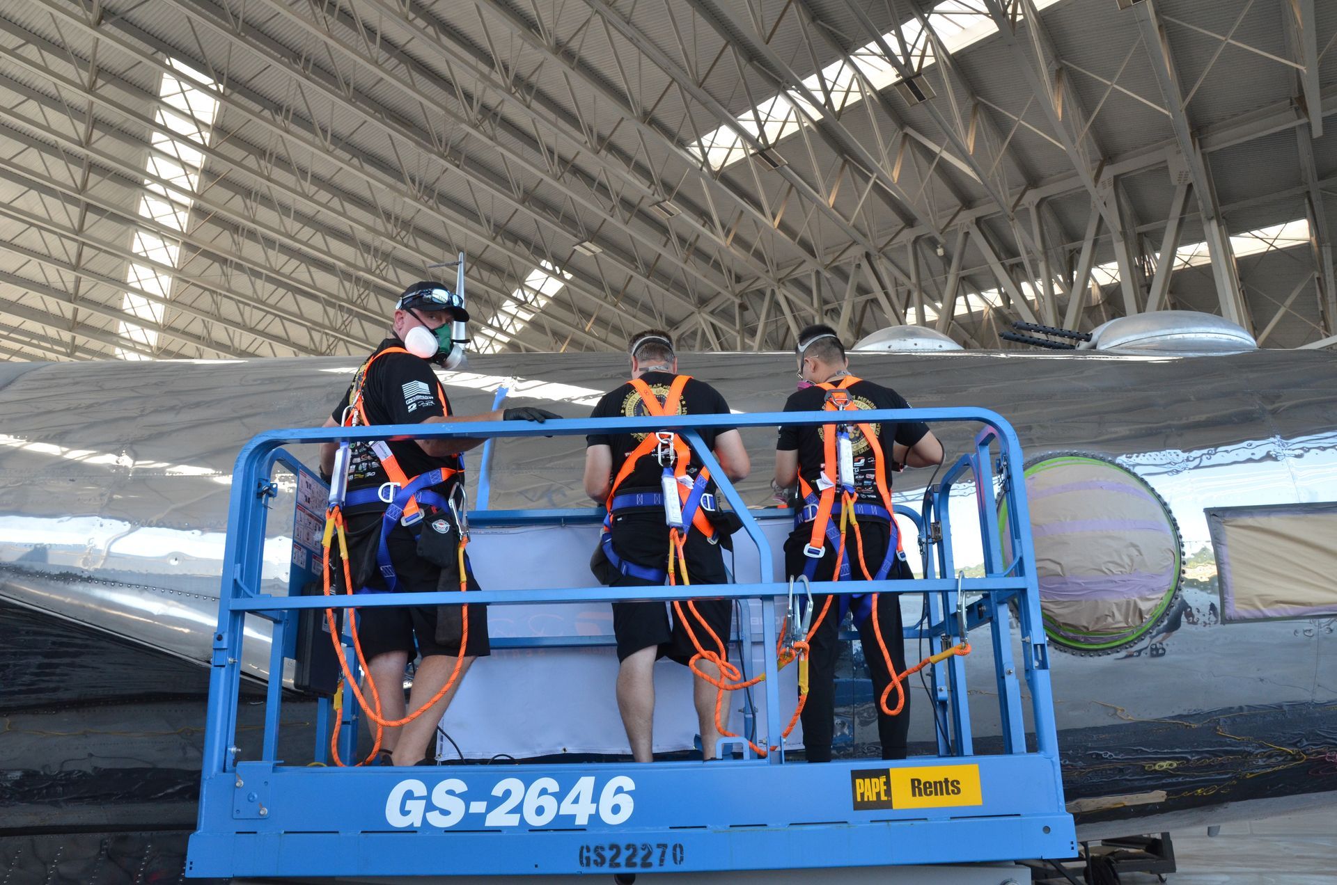 Three people on a blue lift inspecting an airplane wing inside a hangar. They wear safety harnesses.