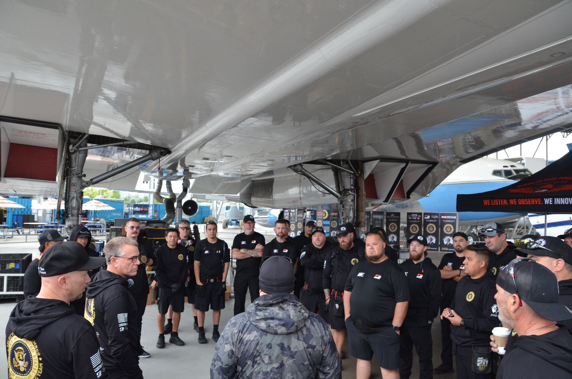 A group of people in black shirts are gathered under a boat, listening to someone speak.