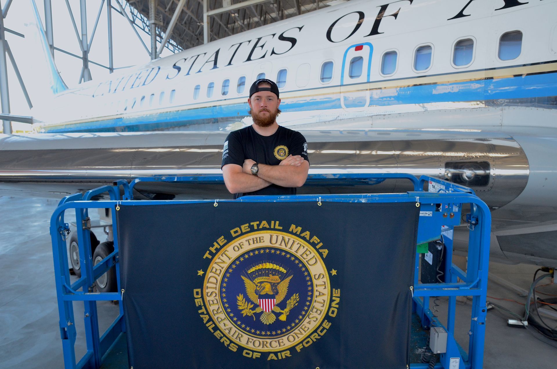 Man stands in front of Air Force One, arms crossed. He is in a blue lift with a banner of 
