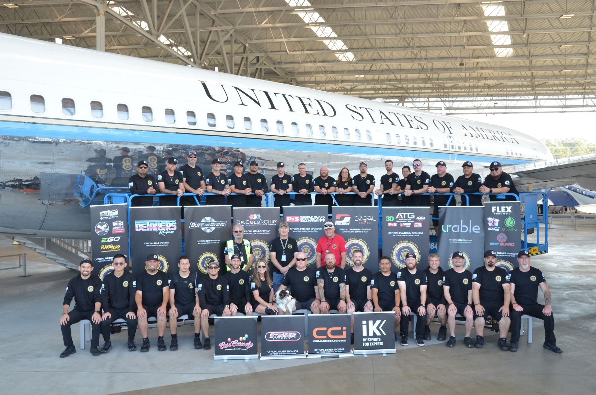 Group photo in front of a plane; people in black shirts, standing and sitting; banners with logos.